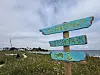 Treskilt med piler og rullestolsymbol står ved en gresskledd strand med sjø i bakgrunnen.