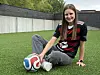 Ung kvinne sitter på kunstgress med en fotball ved siden av seg og en mørk fotballdrakt.