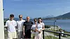 Fem personer står ved et gjerde med fjord, båt og fjell i bakgrunnen.