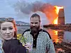 To personer står i forgrunnen foran en stor flamme og røyk ved kysten.