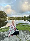 Kvinne sitter på en stein ved en stille innsjø med trær og speilrefleksjoner i vannet.