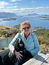 Kvinne med solbriller holder en svart hund foran et fjordlandskap og fjell i solskinn.