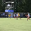 Flere unge spillere i kamp på en fotballbane med publikum langs sidelinjen og et stillas i bakgrunnen.