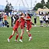 Tre fotballspillere i røde drakter klemmer hverandre på en gressbane under en kamp.