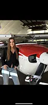 En kvinne står foran et rødt og hvitt småfly inne i en hangar.