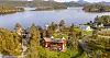 Arkitekttegnet hytte med rød fasade og stor terrasse i et grønt landskap med fjord og fjell i bakgrunnen.