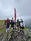 Fire personer står på en steinete fjelltopp under en grå himmel ved et rødt flagg.