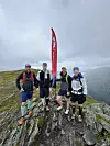 Fire personer står på en steinete fjelltopp under en grå himmel ved et rødt flagg.