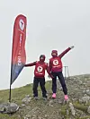 To personer står på en steinete fjelltopp ved et rødt DNT Sunnmøre-flagg i tåke.