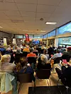 Mange tilhørere sitter tett i en lys sal med store vinduer mens et foredrag holdes foran.
