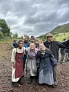En gruppe mennesker i middelalderinspirerte kostymer står tett sammen ute på en gjørmete plass under overskyet himmel.