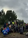 En gruppe mennesker står og sitter samlet på en gresskledd bakketopp under en skyet himmel.