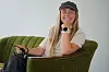 Smilende kvinne i svart caps sitter i en grønn stol og holder et kamera.