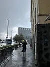 Våt bygatescene med en person som går på regnblankt fortau ved en bygning.