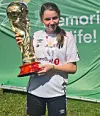 En jente i fotballdrakt holder et stort gullfarget trofé foran en grønn reklamevegg.