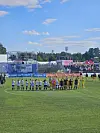 To fotballag står oppstilt på gressbanen foran en fullsatt tribune og bannere.
