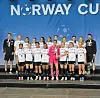 En gruppe unge fotballspillere står på en scene med medaljer, pokal og Norway Cup-bakgrunn.