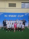 Et jentelag og ledere står samlet foran et blått Norway Cup-banner med en pokal på gressmatte.