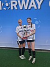 To fotballspillere i hvite drakter står på gresset og holder en pokal foran en blå Norway Cup-vegg.