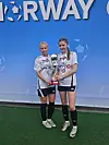 To fotballspillere i hvite drakter står på gresset og holder en pokal foran en blå Norway Cup-vegg.
