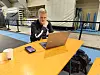 En mann sitter ved et gult bord med laptop, mobiltelefon og kaffekopp i en idrettshall.