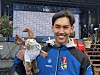 En ung mann i blå jakke holder en medalje og et lite trofé foran en stor resultattavle.