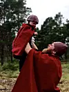 En voksen løfter et barn i røde kapper ute foran trær og himmel.