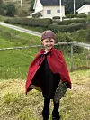 Et barn står ute på gress med rød kappe, mørke klær og lue, foran en grønn bakke og bolighus.