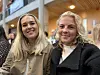 To smilende kvinner står side om side innendørs med varm belysning og trevegger i bakgrunnen.