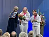 Tre personer står på en scene med blomster og taler under en høytidelig seremoni.