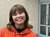 En smilende kvinne i oransje hettegenser står foran en murvegg og en glassdør.