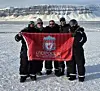 Fem personer i vinterklær holder et rødt Liverpool FC-flagg på snøen foran et fjell.