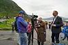 Mennesker står samlet utendørs ved vannet med et kamera på stativ og fjell i bakgrunnen.