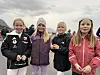 Fire jenter står side om side utendørs i kaldt vær med overskyet himmel i bakgrunnen.