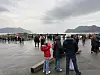 Mange mennesker står samlet på en våt kai ved fjorden under en overskyet himmel.