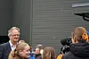 En gruppe mennesker står samlet ute foran en bygning mens en kameramann filmer og en mann snakker.
