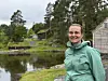 Person i grønn jakke ved en innsjø med skog og bygninger i bakgrunnen.