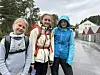 Tre ungdommer med ryggsekker står ved en våt vei i regnvær.