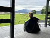Person sitter på en veranda med utsikt mot grønt landskap, vann og fjell.