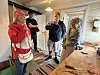 Fire personer står og snakker i et lyst kjøkken med kjøkkenøy, treinnredning og hengende lamper.