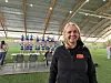 Smilende kvinne foran et gymnastikklag i en lys innendørshall.