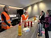 Fire personer i refleksvester står ved et bord med flasker i et innendørs arrangementslokale.