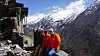 Tre personer i røde jakker sitter ved en steinmur med snødekte fjell og dal bak.
