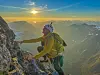 En klatrer i gul jakke klatrer på en bratt fjellrygg med solnedgang og fjell i bakgrunnen.