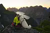 Person med hund sitter på en fjellknaus og ser mot fjell og fjord i solnedgang.