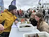 Folk står ved en stand med esker og beholdere ved kaia, med båter og festivalområde i bakgrunnen.