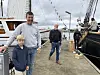 To menn og en gutt står på en kai ved siden av en stor trebåt og andre båter med flagg i havna.