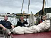 Tre menn sitter på dekk i en båt med tau og seilduk om bord, ved en havn med andre båter i bakgrunnen.