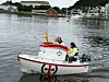 En liten hvit båt med røde detaljer og Elias-merke seiler med to personer om bord på rolig vann i en havn.