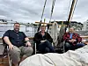 Tre menn sitter på dekk i en seilbåt ved en havn med båter og bygninger i bakgrunnen.